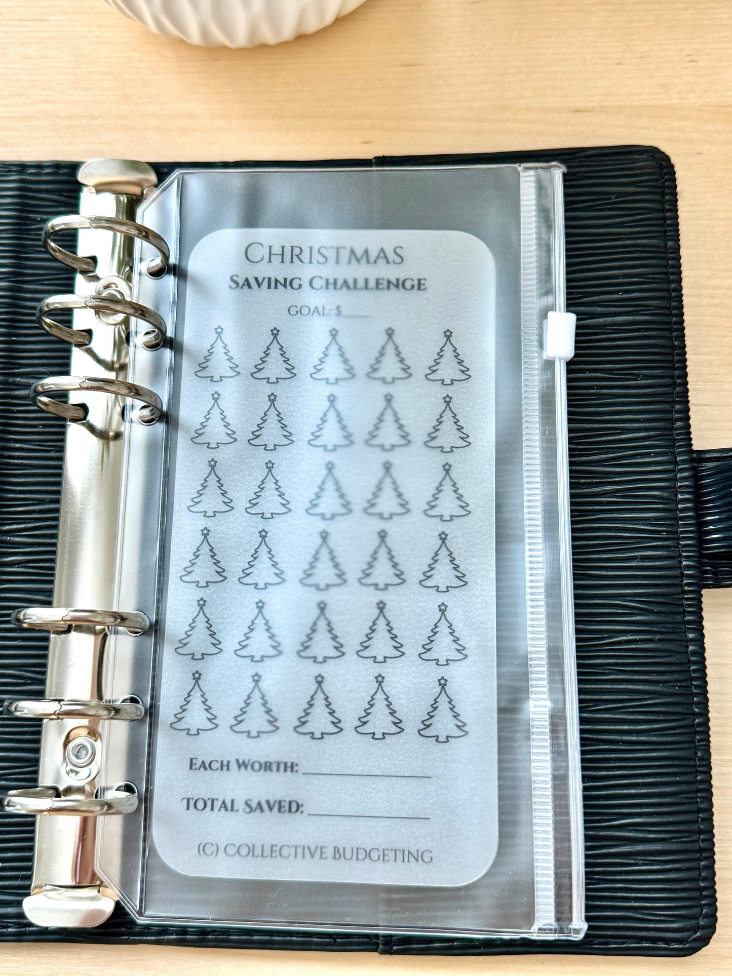 A clear plastic sheet with a Christmas saving challenge tracker. The sheet has a black outline of 36 Christmas trees. The text on the sheet reads 'Christmas Saving Challenge', 'Goals', 'Each Worth', 'Total Saved', and '(C) Collective Budgeting'.