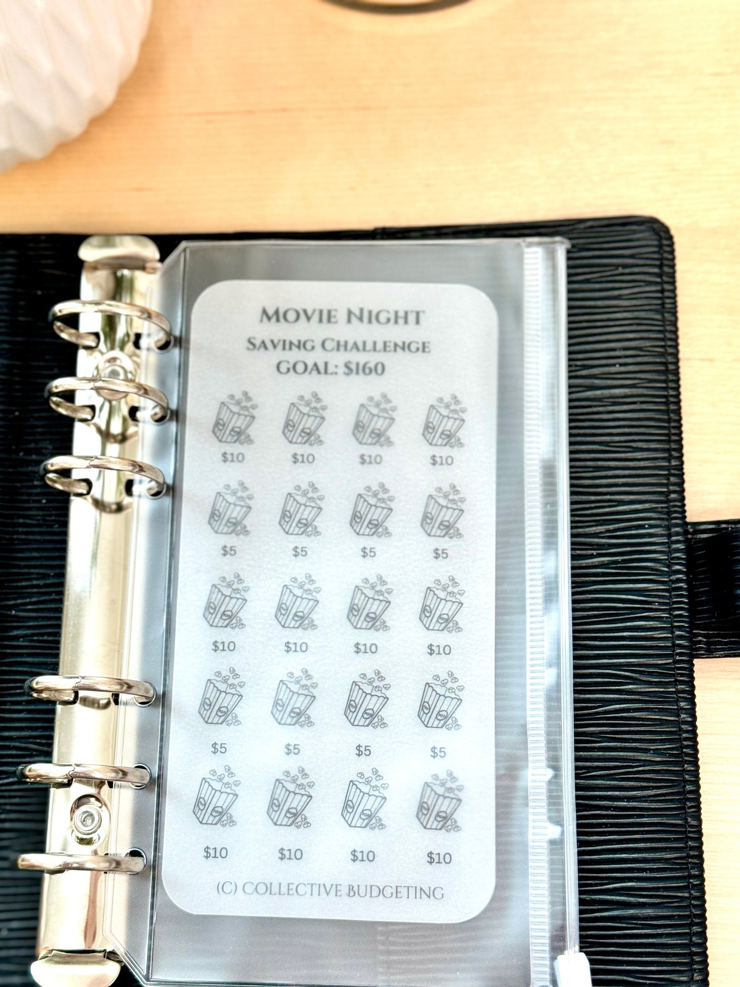 A clear plastic page with a white background and black text. The text reads 'Movie Night Saving Challenge Goal: $160'. The page is divided into 20 squares, each with a black and white illustration of a popcorn bucket. The squares are labeled with the amounts '$10' and '$5'. The bottom of the page reads '(C) Collective Budgeting'.