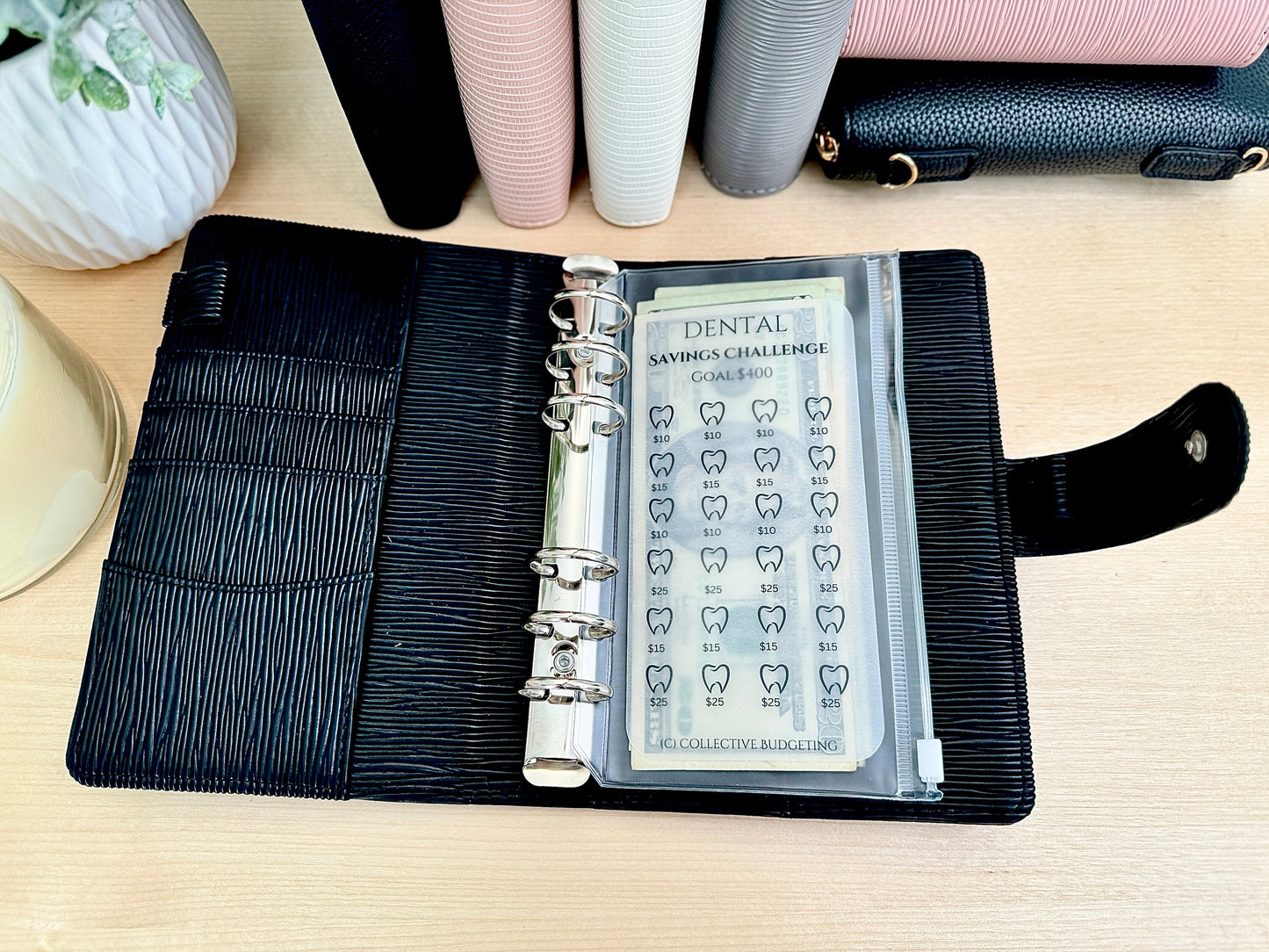 A black leather planner with a clear plastic pocket containing a dental savings challenge tracker. The tracker has a goal of $400 and features a grid of teeth with dollar amounts next to each tooth. The tracker is labeled 'Dental Savings Challenge' and 'Collective Budgeting'.