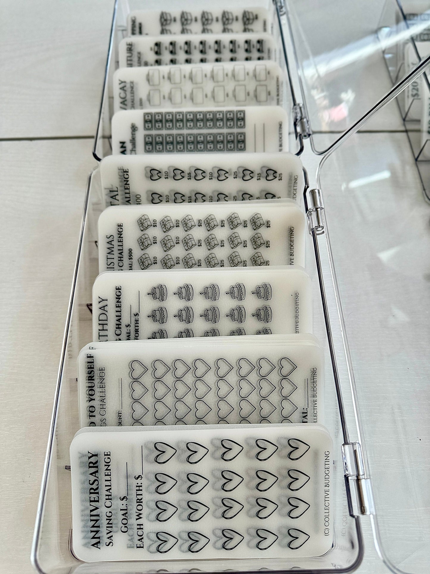 A clear plastic storage box containing eight transparent cards with black ink. Each card has a different savings challenge theme, including anniversary, birthday, Christmas, and vacation. The cards are labeled with the challenge name, goal amount, and value of each item. The cards are designed for collective budgeting.