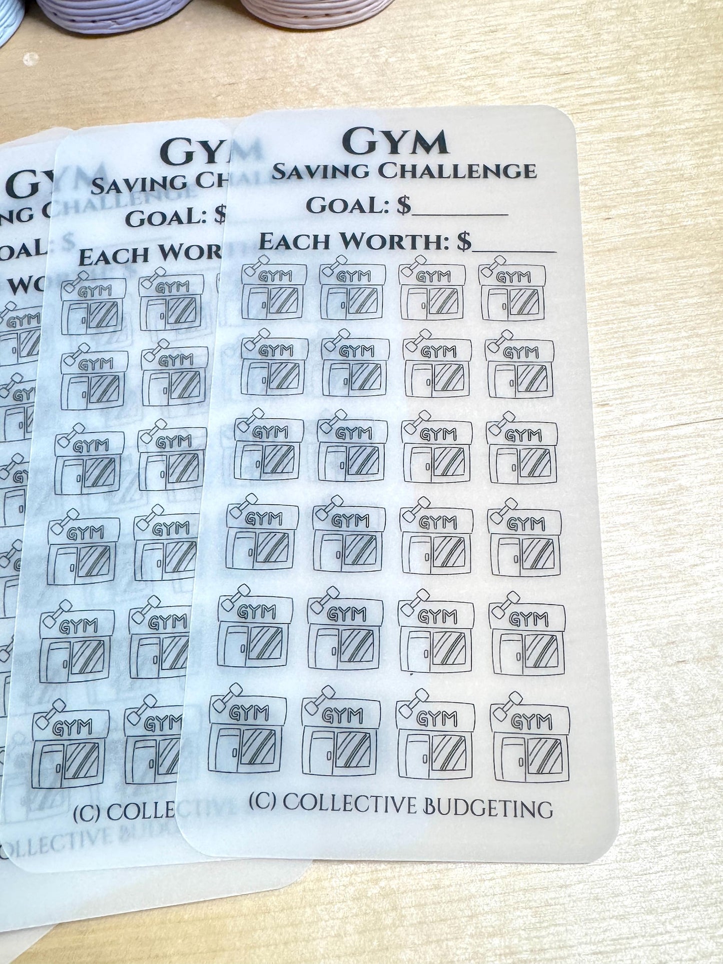 A close-up of a clear sheet with a black and white gym saving challenge tracker. The sheet has a title 'Gym Saving Challenge' with two blank lines for the goal amount and the amount each gym visit is worth. The sheet has 48 black and white gym icons with the word 'Gym' written on each icon. The bottom of the sheet has the text '(C) Collective Budgeting'.