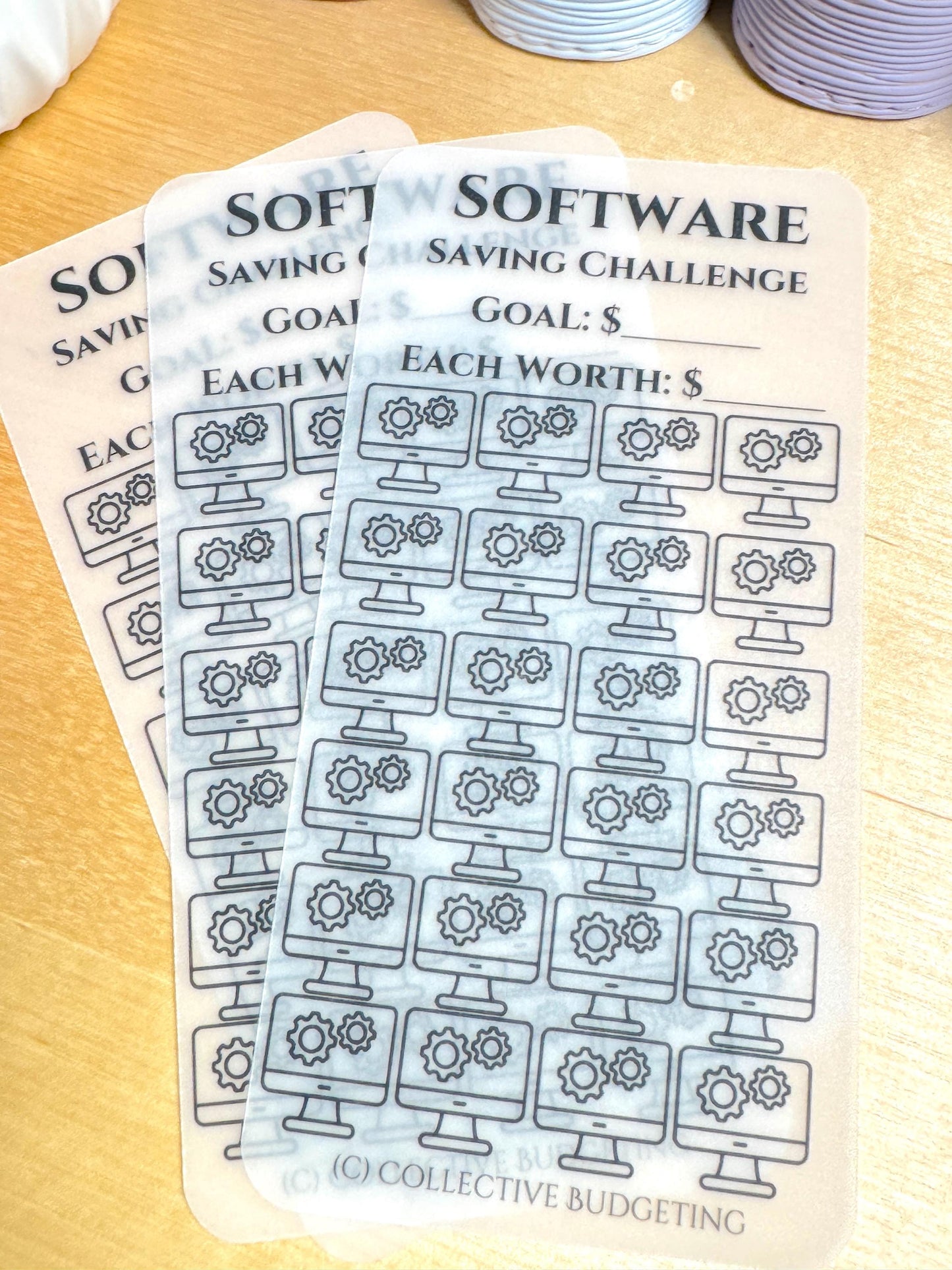 Two transparent sheets with black text and graphics. The top sheet has the text 'SOFTWARE SAVING CHALLENGE' and the text 'GOAL: $' and 'EACH WORTH: $'. The bottom sheet has the text 'SOFTWARE SAVING CHALLENGE' and the text 'GOAL: $' and 'EACH WORTH: $'. Both sheets have a grid of 48 computer monitor icons with three gears inside each monitor. The bottom sheet has the text '(C) COLLECTIVE BUDGETING' at the bottom.