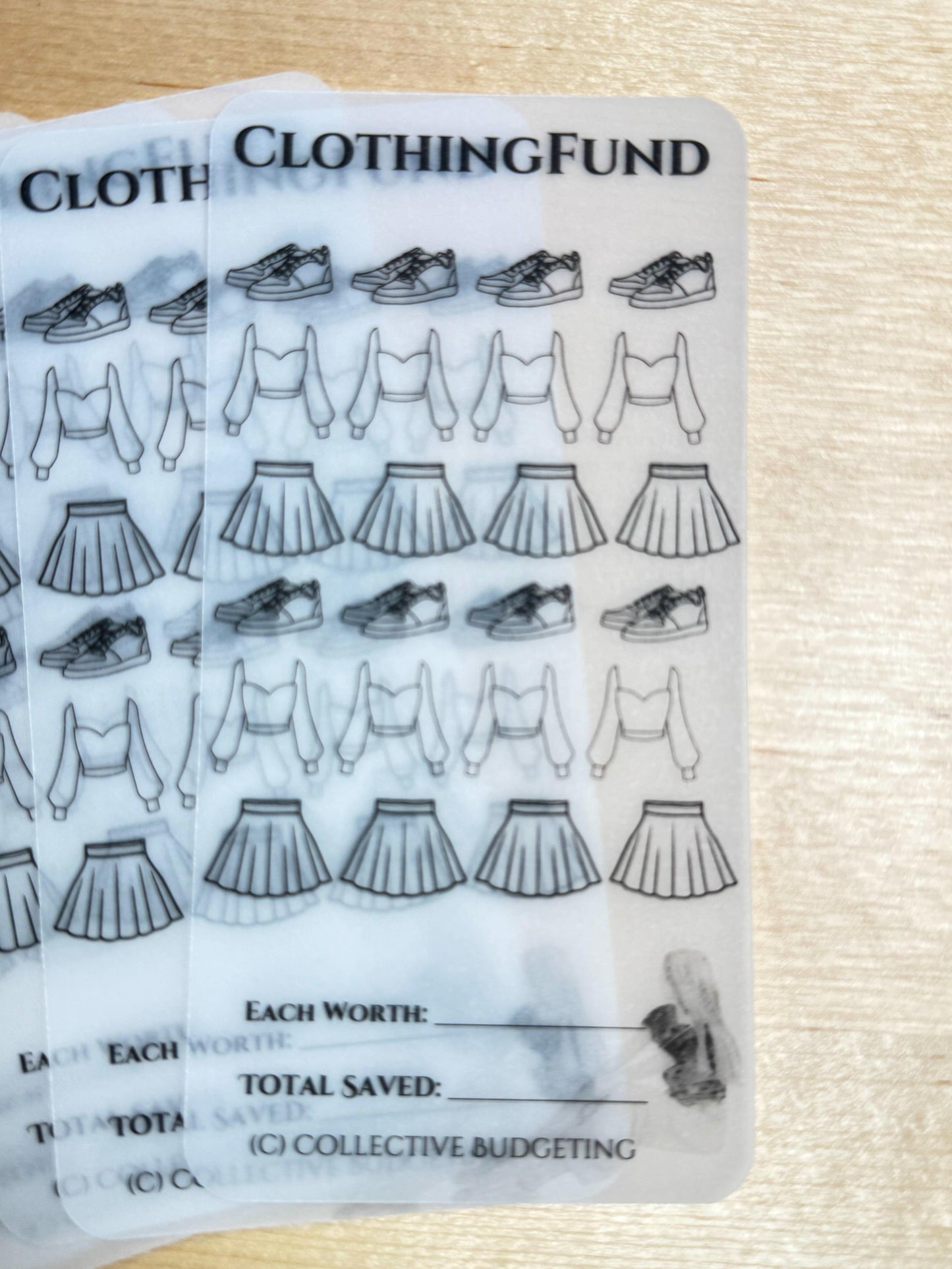 A clear plastic sheet with black line drawings of clothing items, including sneakers, tops, and skirts. The sheet has the text 'CLOTHINGFUND' at the top and the text 'EACH WORTH:' and 'TOTAL SAVED:' at the bottom. The text '(C) COLLECTIVE BUDGETING' is also at the bottom.