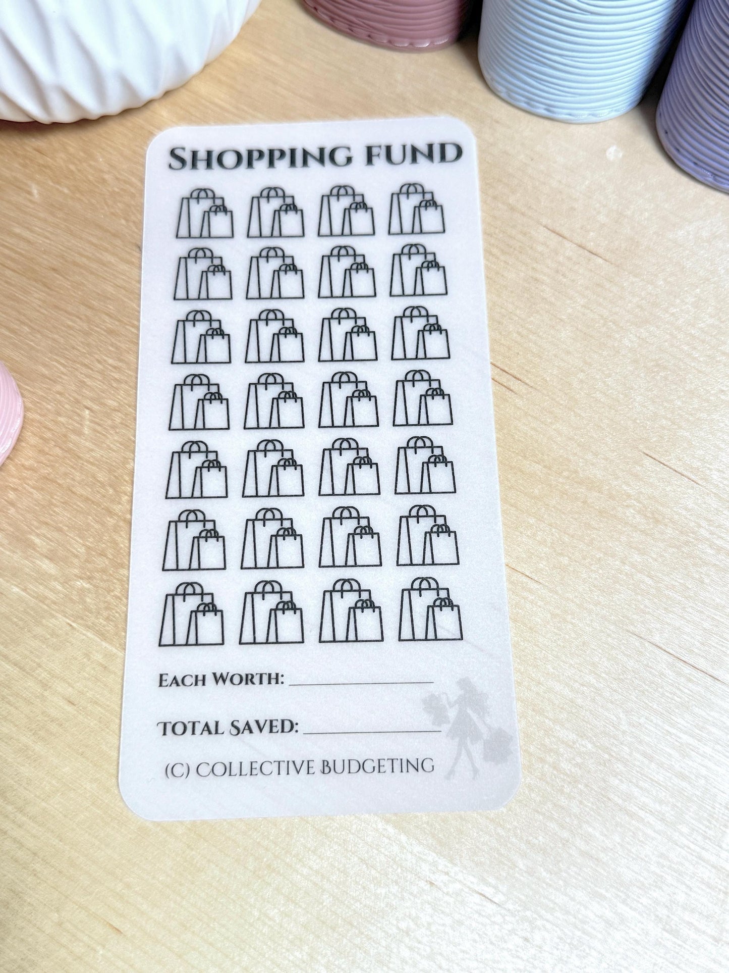 A white sticker sheet with black shopping bag icons. The sheet has the text 'SHOPPING FUND' at the top. There are 30 shopping bag icons in total. The sheet has the text 'EACH WORTH:' and 'TOTAL SAVED:' at the bottom. The sheet also has the text '(C) COLLECTIVE BUDGETING' at the bottom.