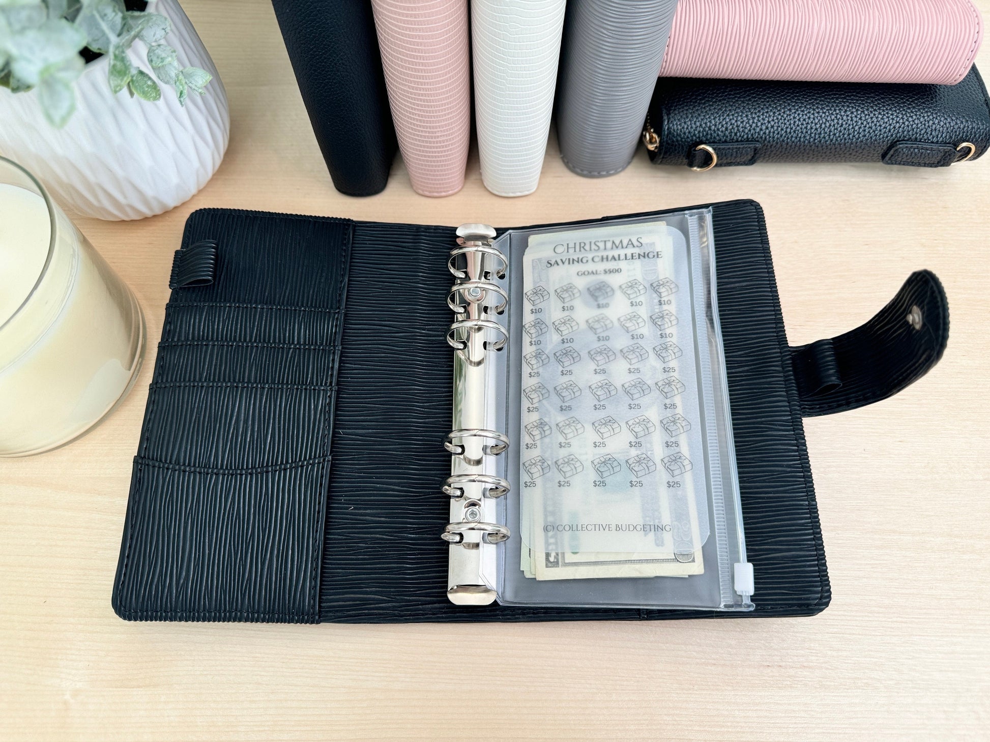 A black leather planner with a silver ring binder. The planner is open to reveal a clear plastic pocket with a Christmas saving challenge sheet inside. The sheet has a grid of boxes with dollar amounts listed in each box. The sheet also has the text 'Christmas Saving Challenge' and 'Collective Budgeting'.