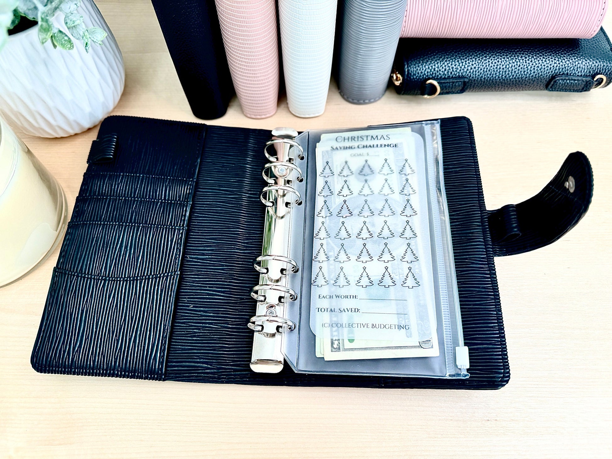 A black leather planner with silver rings and a clear plastic pocket containing a Christmas savings challenge tracker. The tracker has a list of 25 Christmas trees with a space to write the amount saved for each tree. The text on the tracker reads 'Christmas Saving Challenge', 'Each Worth', 'Total Saved', and '#collectivebudgeting'.