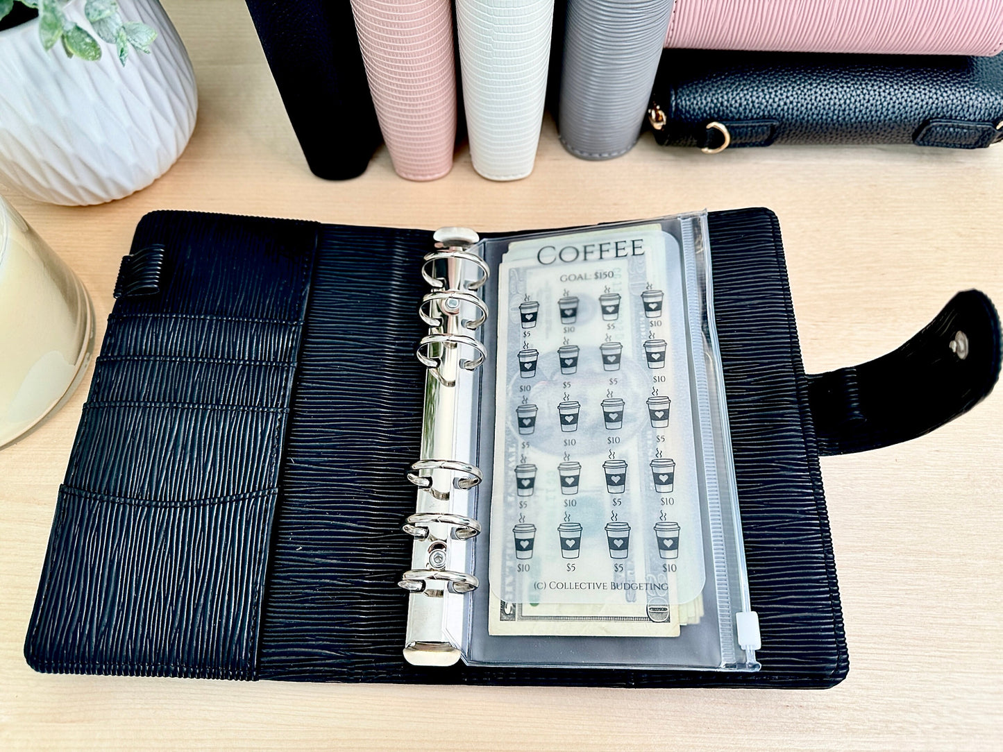 A black leather planner with silver rings and a clear plastic pocket containing a coffee sticker sheet. The sticker sheet has a coffee cup icon and the text 'COFFEE GOAL RING' and '50 COLLECTIVE BUDGETING'.