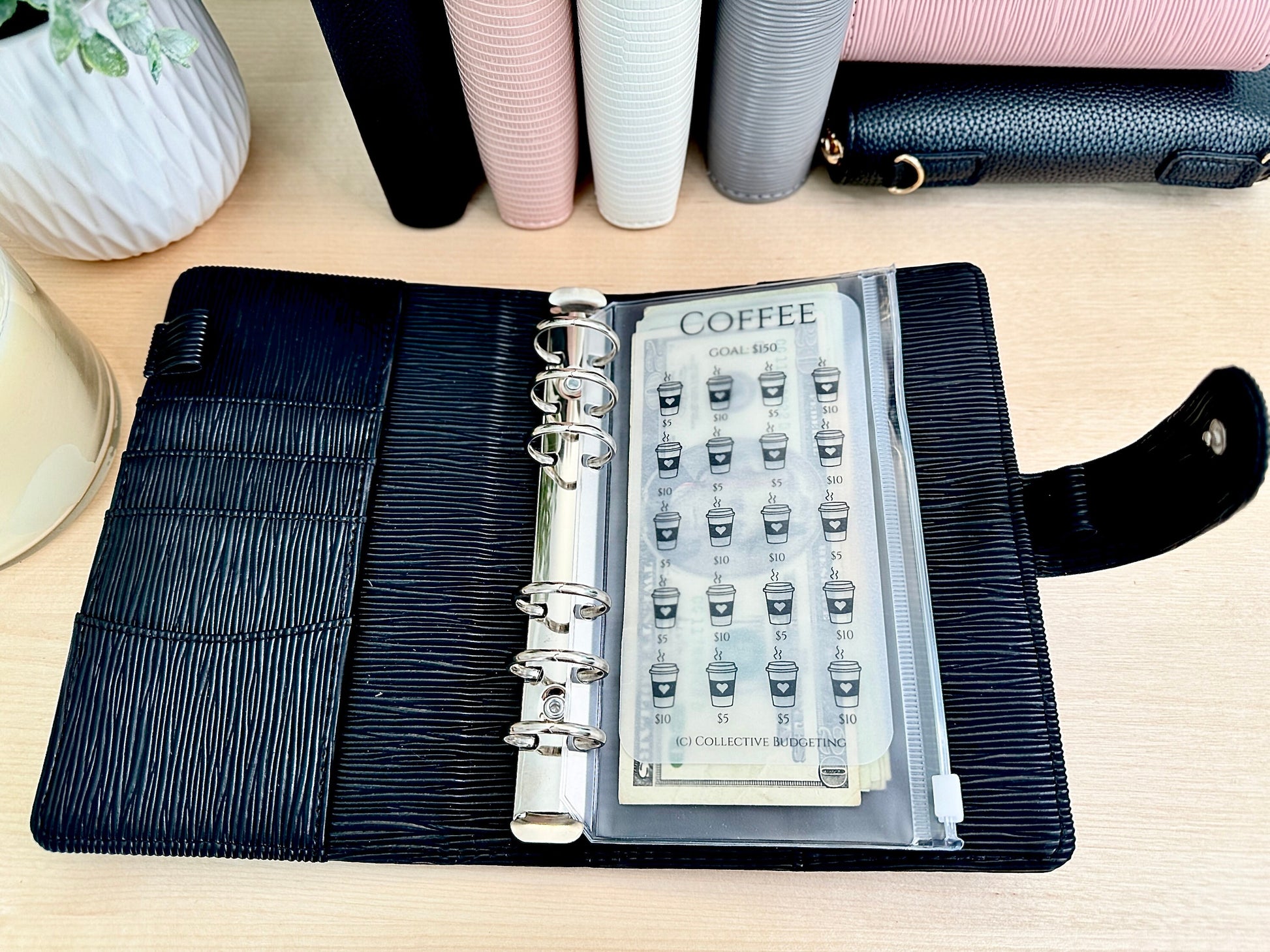 A black leather planner with silver rings and a clear plastic pocket containing a coffee sticker sheet. The sticker sheet has a coffee cup icon and the text 'COFFEE GOAL RING' and '50 COLLECTIVE BUDGETING'.