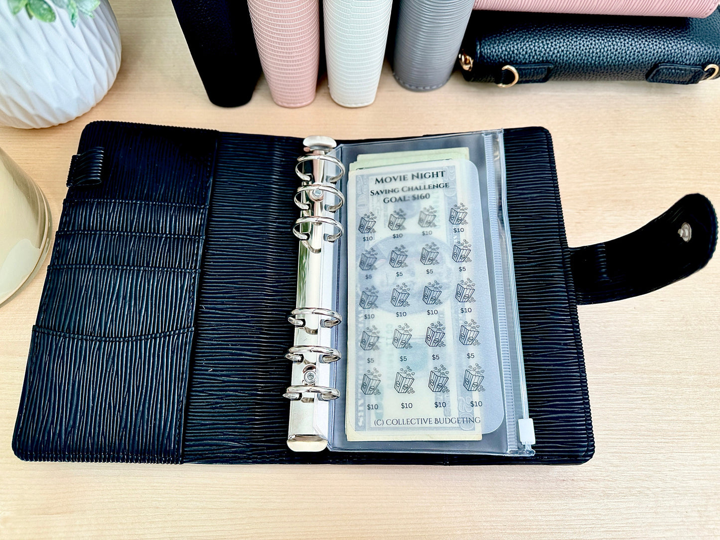 A black leather planner with a clear plastic pocket containing a movie night savings challenge sheet. The sheet has a goal of $160 and includes 16 boxes with a movie ticket icon and a dollar amount of $10 in each box. The text on the sheet reads 'Movie Night Saving Challenge Goal $160' and 'Collective Budgeting'.