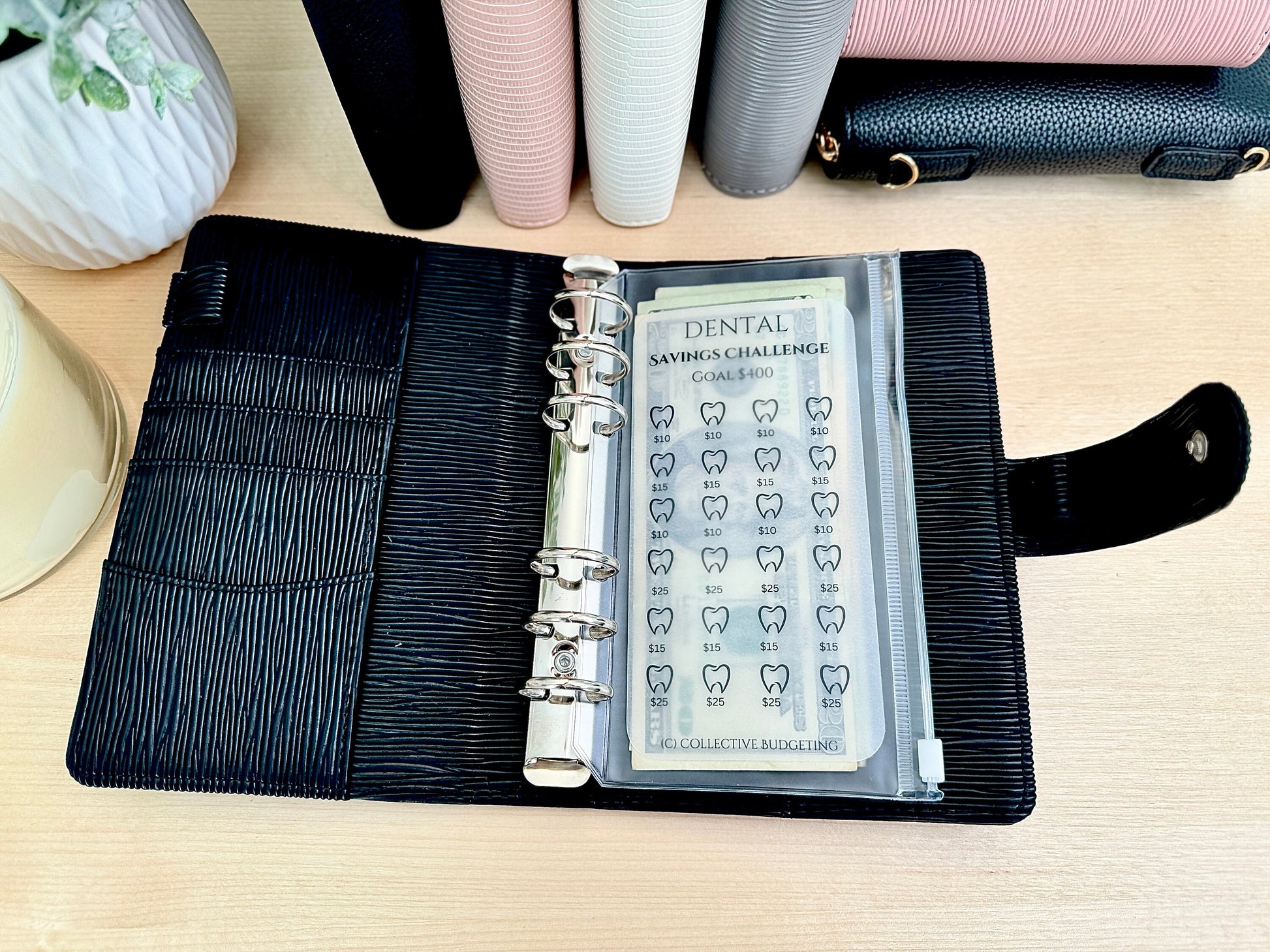 A black leather planner with a clear plastic pocket containing a dental savings challenge tracker. The tracker has a goal of $400 and features a grid of teeth with dollar amounts next to each tooth. The tracker is labeled 'Dental Savings Challenge' and 'Collective Budgeting'.