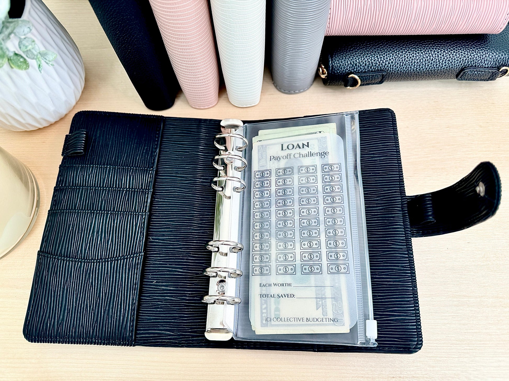 A black leather planner with a silver ring binder. The planner is open and a clear plastic pocket is visible with a sheet of paper inside. The paper has the text 'LOAN Payoff Challenge' and a grid of dollar signs. The text 'Each worth:', 'Total Saved:', and 'Collective Budgeting' is also visible on the sheet.