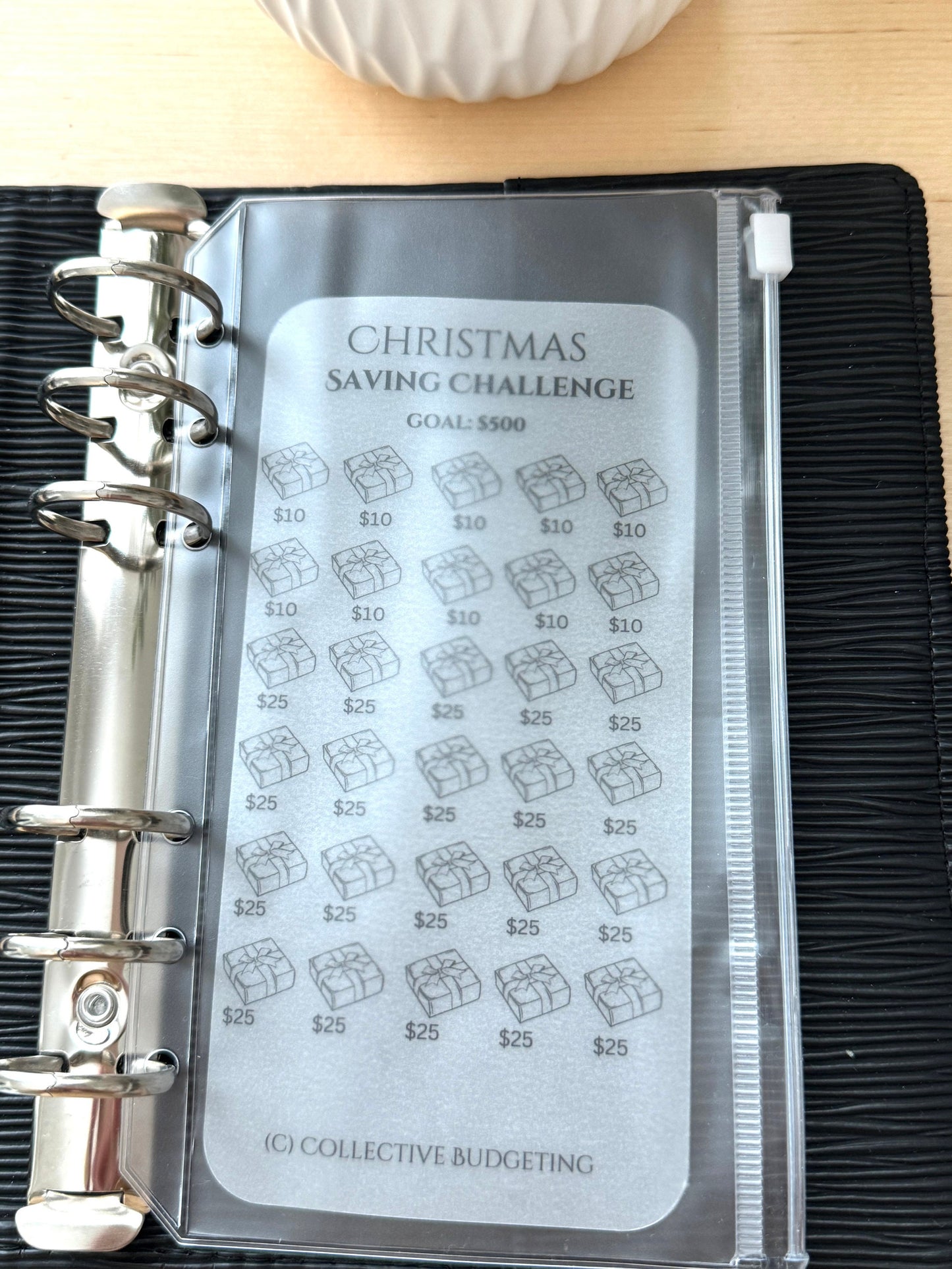 A gray and white Christmas saving challenge tracker with a zipper closure. The tracker has a goal of $5500 and features a grid of 25 gift boxes with a dollar amount of $10 or $25. The text 'CHRISTMAS SAVING CHALLENGE' and '(C) COLLECTIVE BUDGETING' is printed on the tracker.