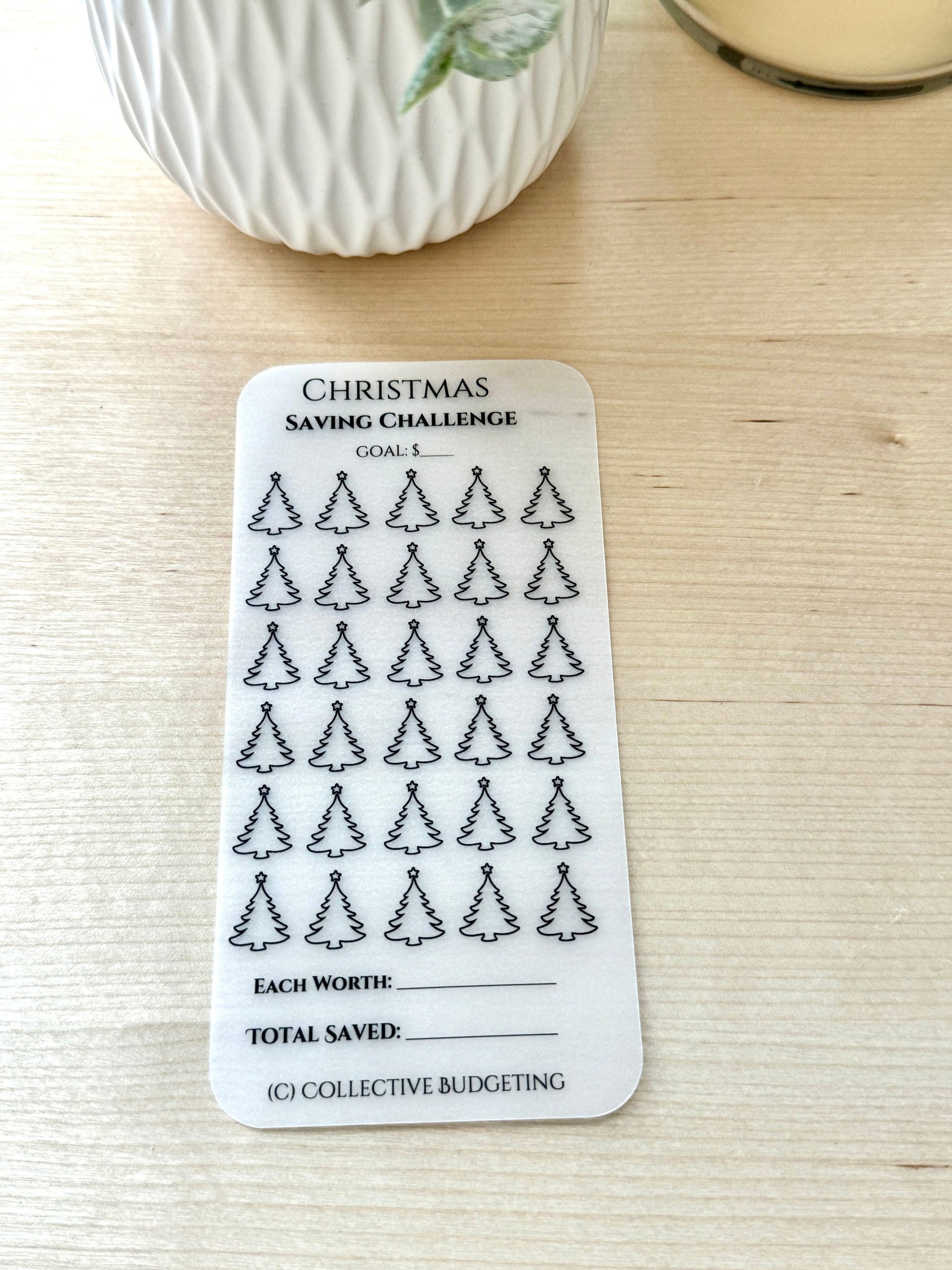 A white card with black text and outlines of Christmas trees. The card is titled 'Christmas Saving Challenge.' The card has a section for a goal amount, a section for the value of each tree, and a section for the total saved. The text at the bottom of the card reads '(C) COLLECTIVE BUDGETING.'