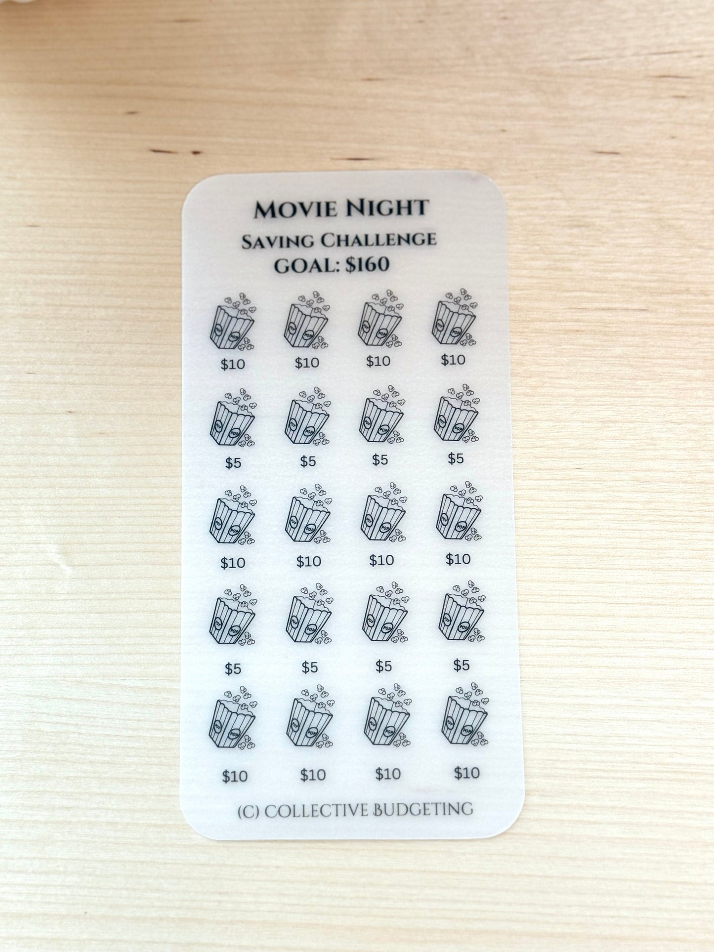 A clear laminated vellum card with black text and images. The text reads 'Movie Night Saving Challenge Goal: $160'. The sticker has a grid of 20 images of popcorn buckets with dollar amounts next to each image. The dollar amounts are $5, $10, and $10. The bottom of the sticker has the text '(C) Collective Budgeting'.