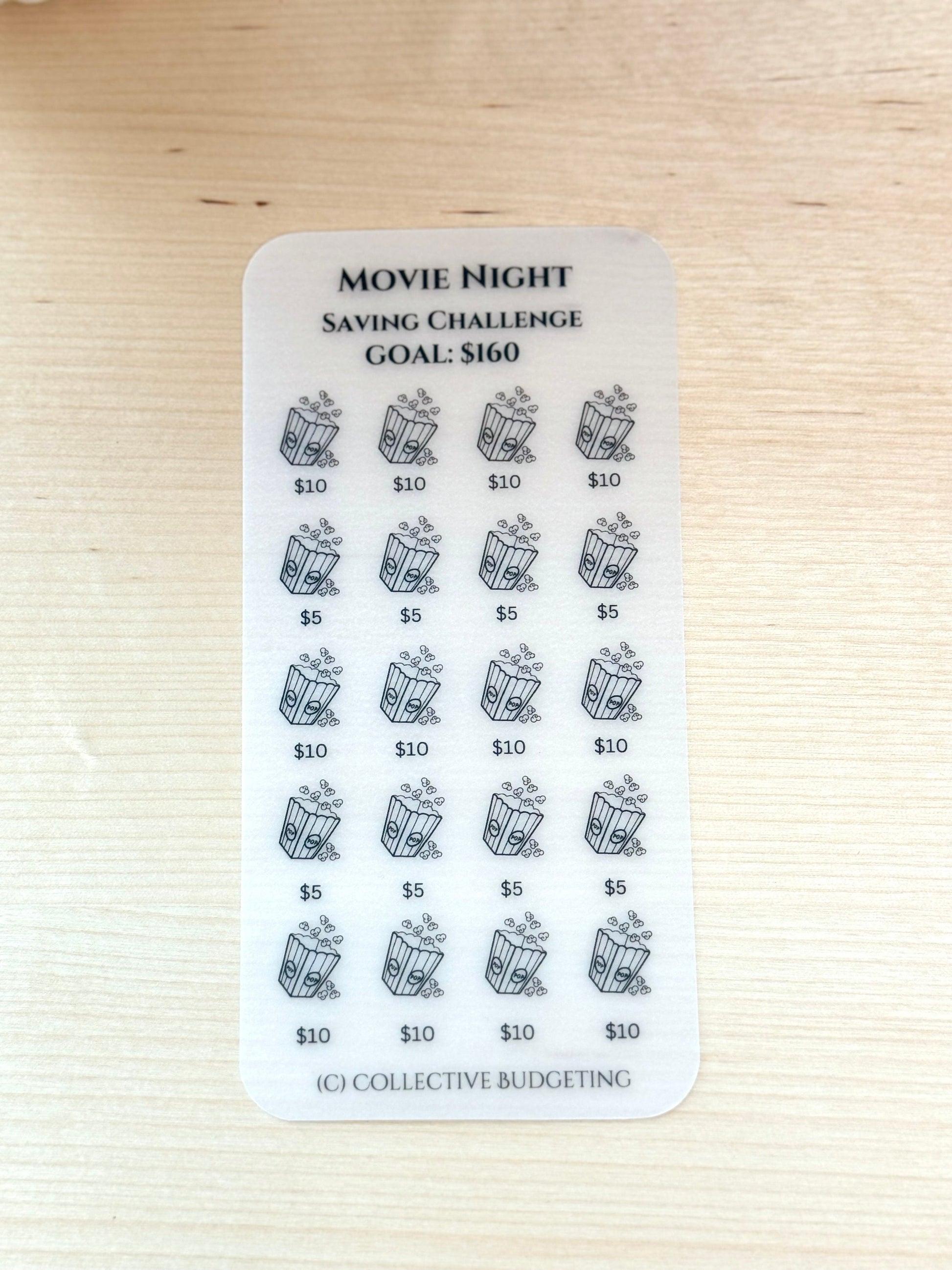 A clear laminated vellum card with black text and images. The text reads 'Movie Night Saving Challenge Goal: $160'. The sticker has a grid of 20 images of popcorn buckets with dollar amounts next to each image. The dollar amounts are $5, $10, and $10. The bottom of the sticker has the text '(C) Collective Budgeting'.