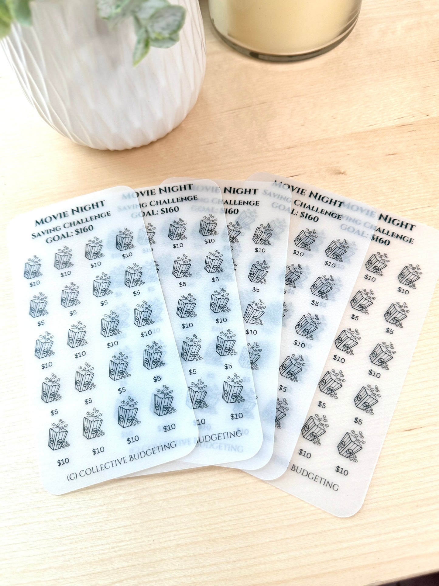 Five transparent sheets with a movie night savings challenge theme. Each sheet has a grid of 30 popcorn buckets with the values $5 and $10 printed on them. The sheets are stacked on top of each other and the text 'Movie Night Saving Challenge Goal: $160' is printed on each sheet. The text '(C) Collective Budgeting' is printed on the bottom of one of the sheets.