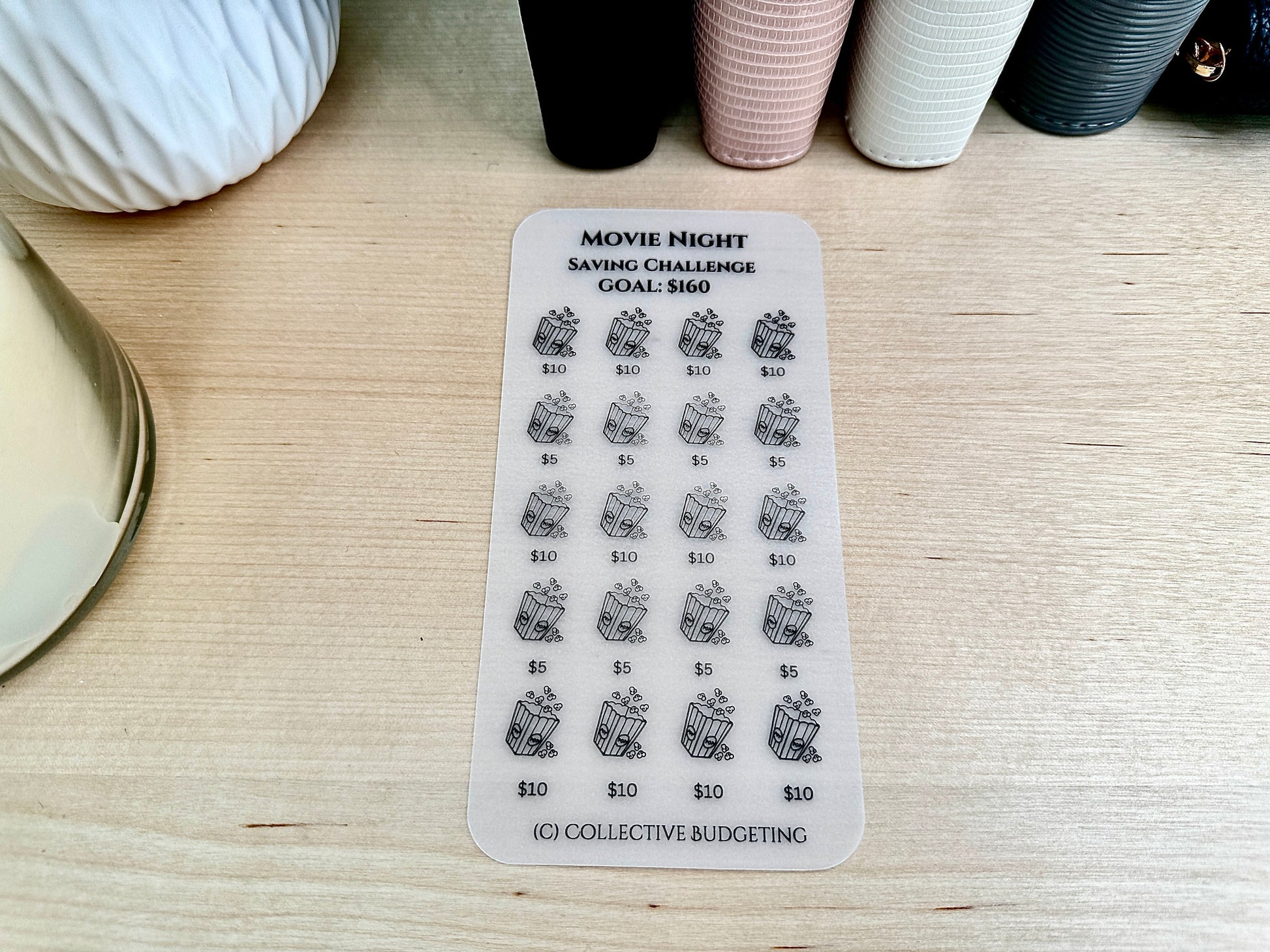 A clear laminated vellum card with black text and images. The text reads 'Movie Night Saving Challenge Goal: $160'. The sticker has a grid of 20 images of popcorn buckets with dollar amounts next to each image. The dollar amounts are $5, $10, and $10. The bottom of the sticker has the text '(C) Collective Budgeting'.