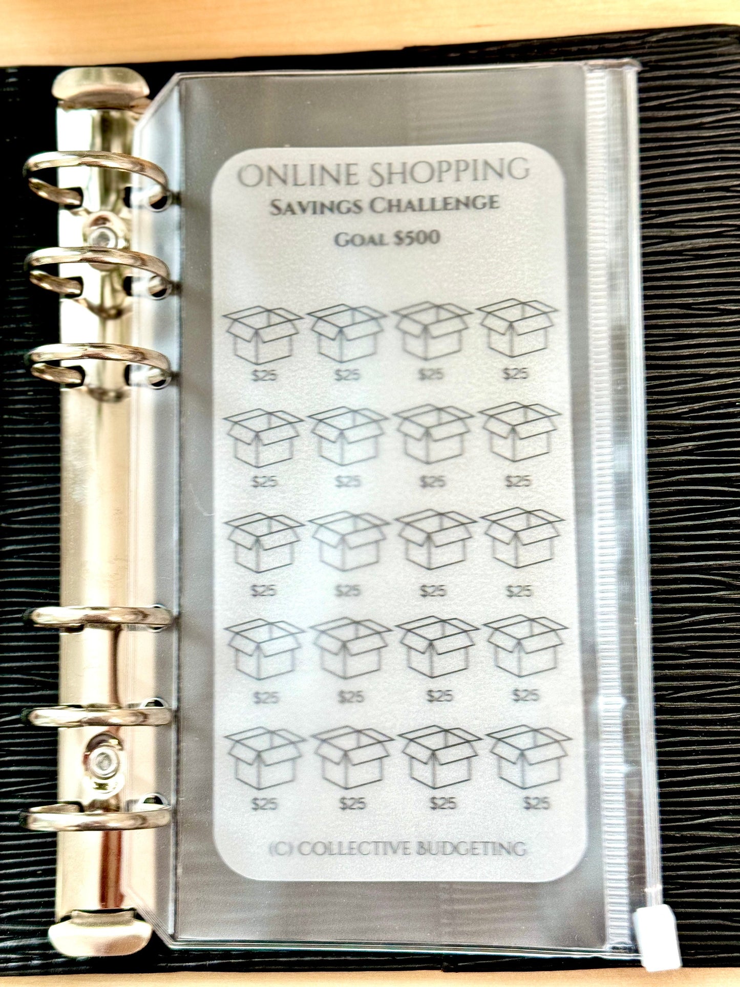 A white plastic sheet with the text 'ONLINE SHOPPING SAVINGS CHALLENGE GOAL $500' and 20 boxes with the text '$25' in each box. The sheet is in a clear plastic binder with silver rings. The text '(C) COLLECTIVE BUDGETING' is at the bottom of the sheet.