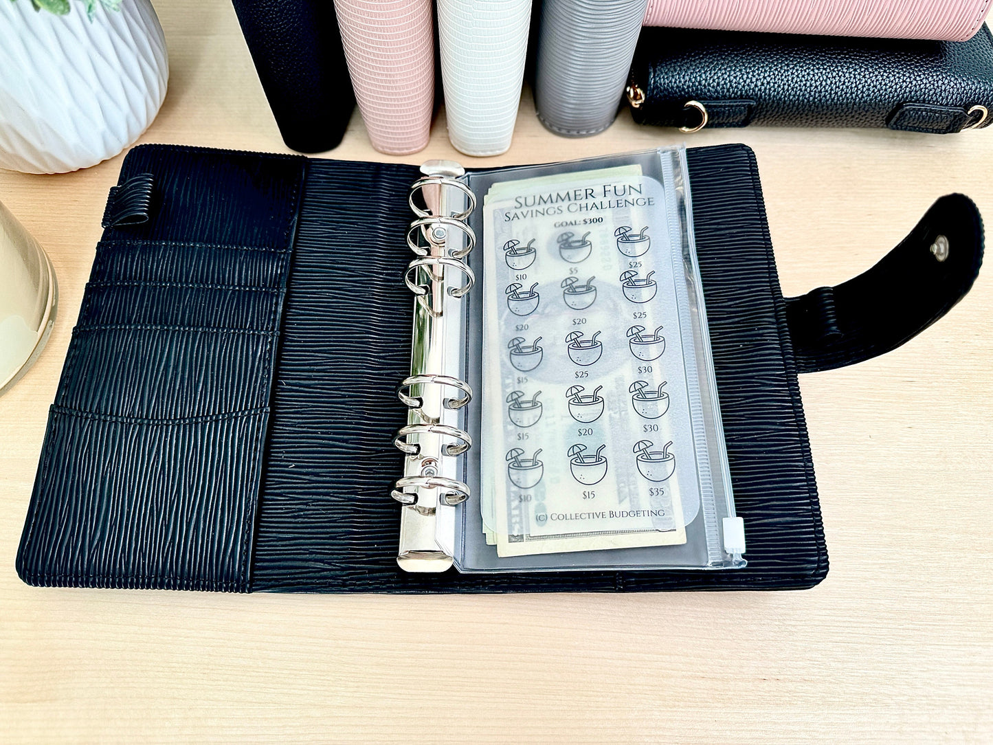 A black leather planner with a clear plastic pocket containing a savings challenge tracker. The tracker is titled 'Summer Fun Savings Challenge' with a goal of $300. The tracker has 15 rows of coconut drink icons with dollar amounts ranging from $5 to $15. The tracker is labeled 'Collective Budgeting' at the bottom.