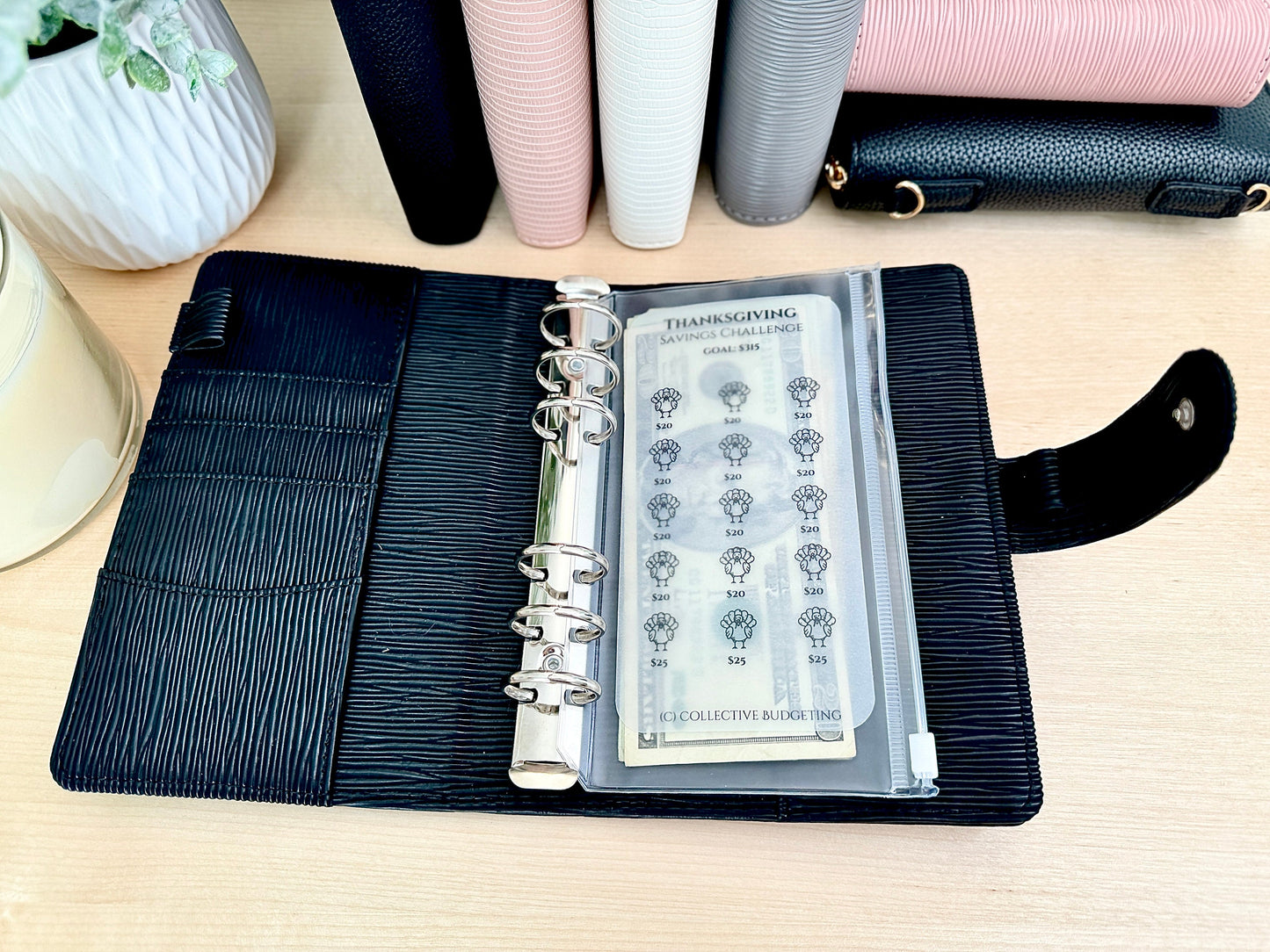 A black leather bound planner with a clear plastic pocket containing a Thanksgiving savings challenge sheet. The sheet has a turkey graphic and lists savings goals from $5 to $25. The text on the sheet reads 'Thanksgiving Savings Challenge Goals $5 - $25 Collective Budgeting'.