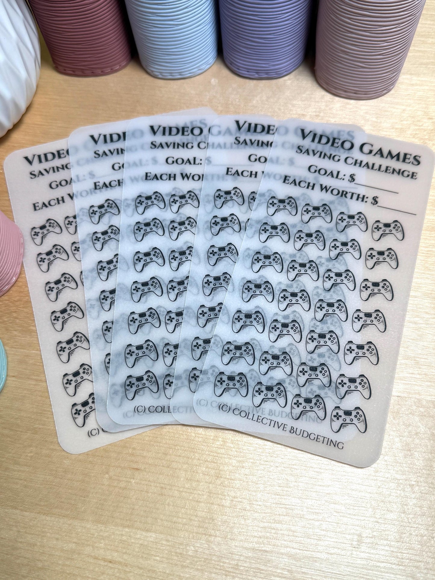 Four sheets of clear plastic stickers with black video game controller icons. The stickers are labeled 'Video Games Saving Challenge' and 'Goal: $ Each Worth: $'. The stickers are designed for a budgeting or savings tracker.