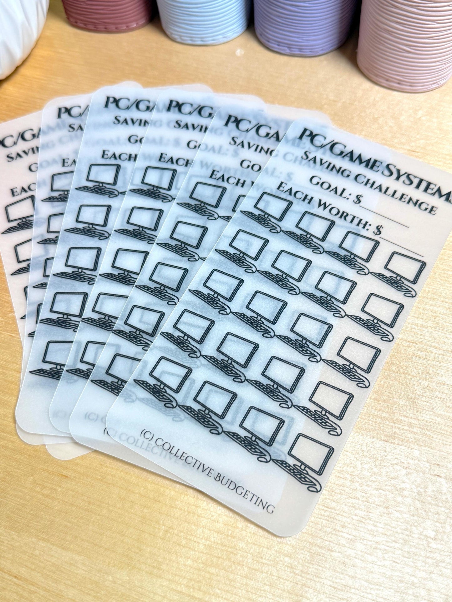 A stack of six transparent sheets with a black and white computer icon repeated across each sheet. The sheets are labeled 'PC/GAME SYSTEM SAVING CHALLENGE GOAL: $ EACH WORTH: $ (C) COLLECTIVE BUDGETING'.