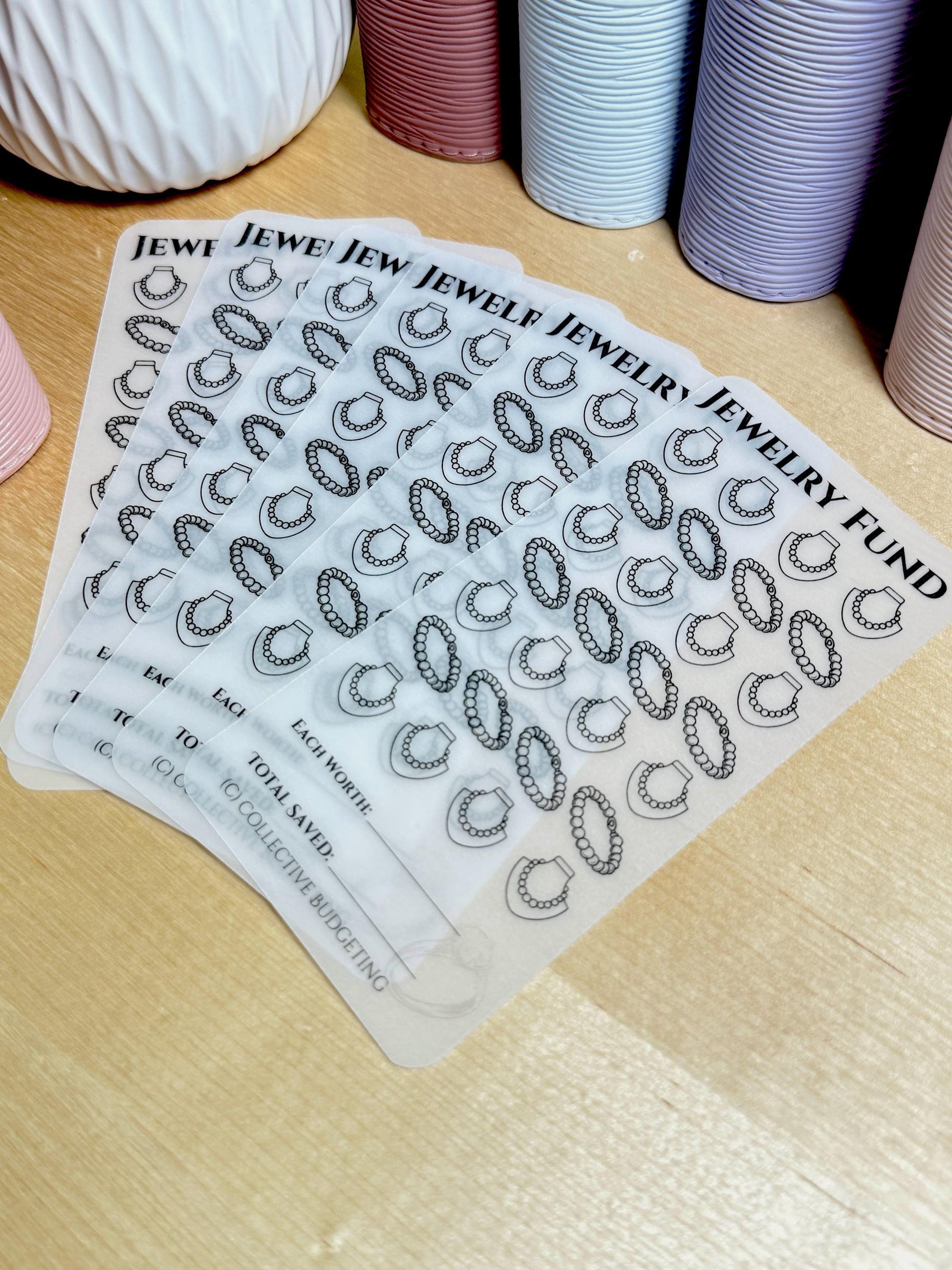 A stack o Five transparent sheets with a black and white design of necklaces. The sheets have the text 'JEWELRY FUND', 'EACH WORTH', 'TOTAL SAVED', and '(C) COLLECTIVE BUDGETING'.