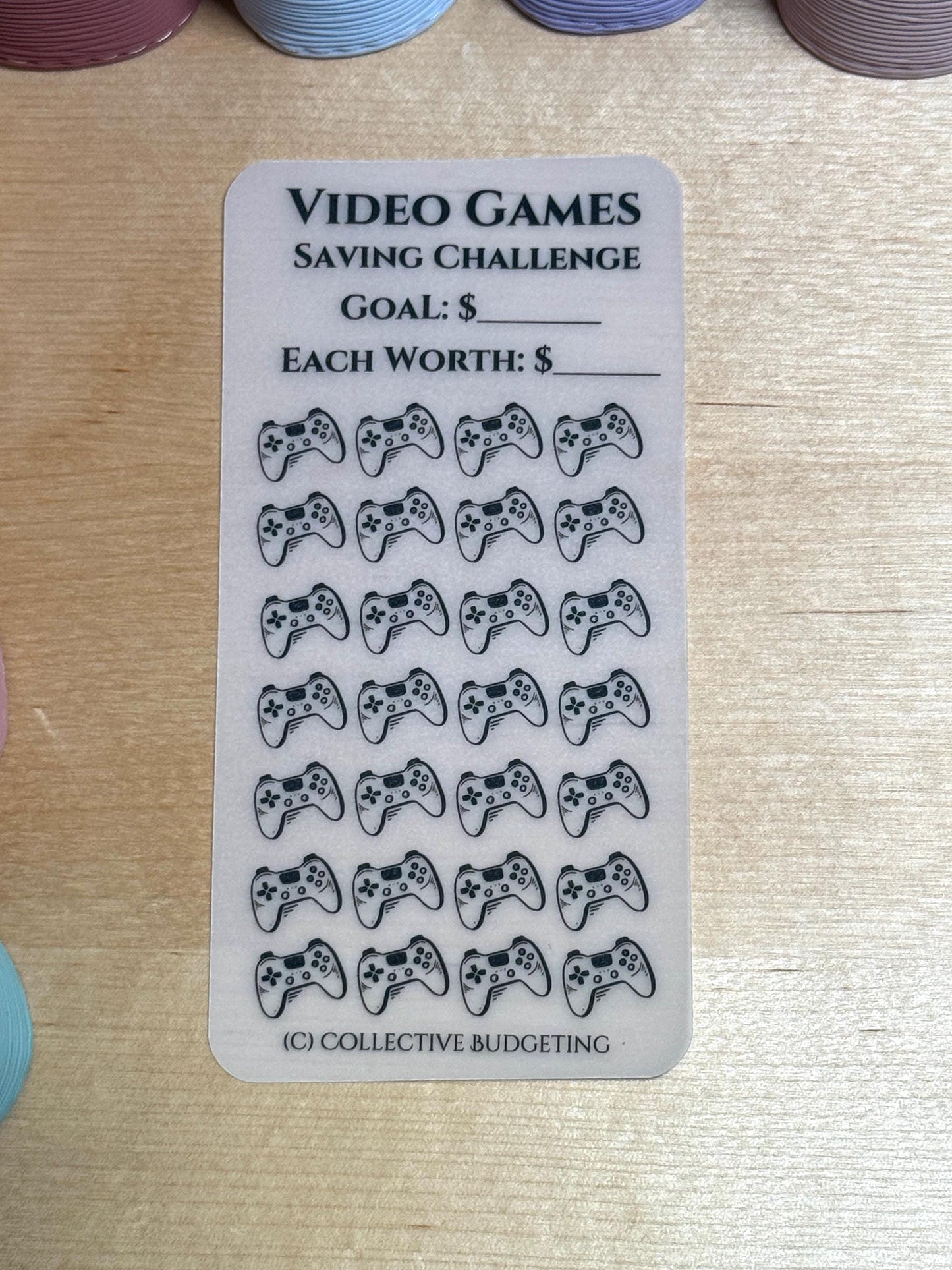 A transparent sticker sheet with a video game saving challenge theme. The sheet has a title that reads 'Video Games Saving Challenge.' There are two blank lines for writing in a dollar amount for the goal and the amount each sticker is worth. The sheet has 25 black and white video game controller icons.