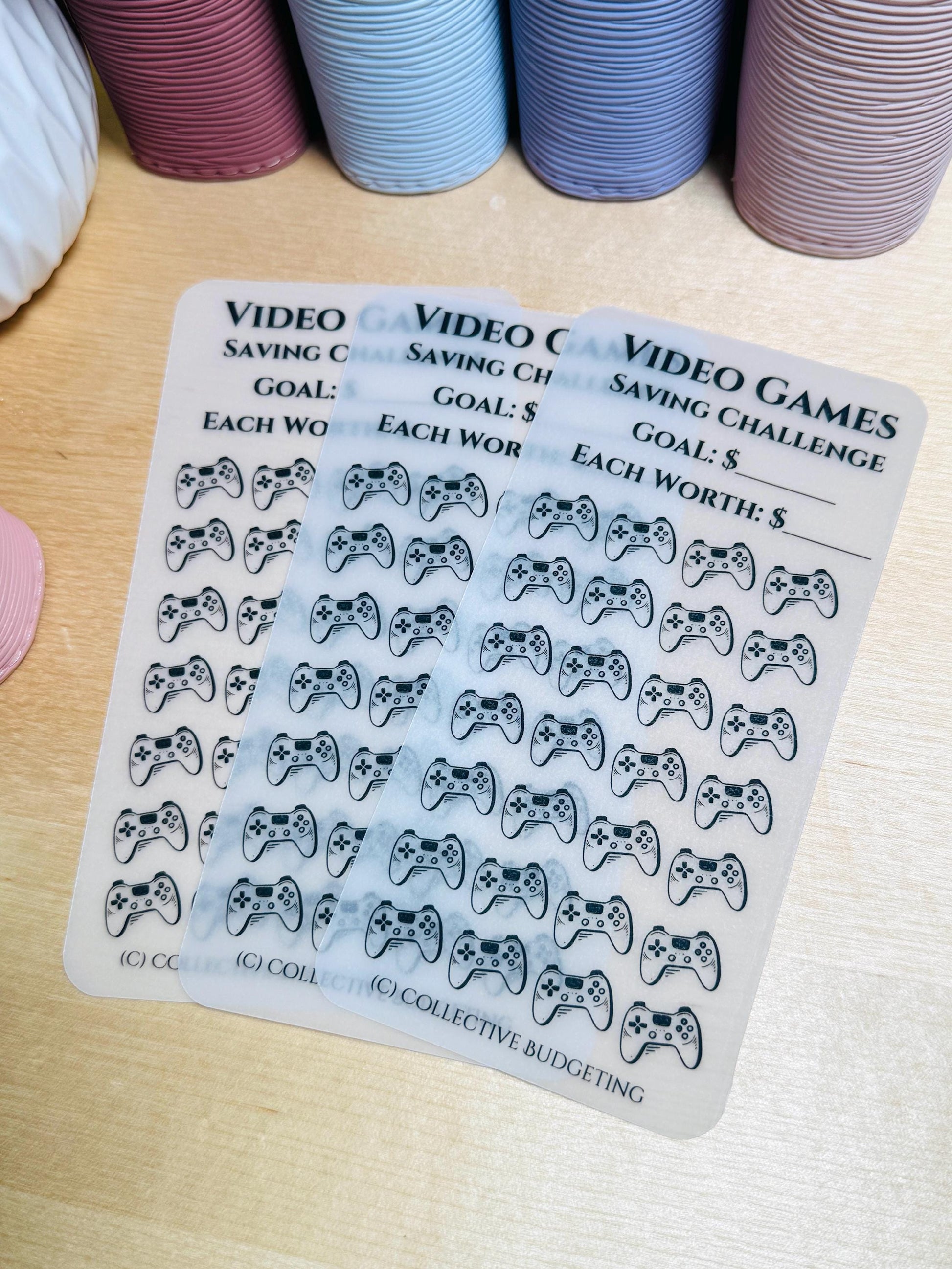 Three white sheets with black video game controller icons. The top sheet has the text 'VIDEO GAMES SAVING CHALLENGE GOAL: $ EACH WORTH: $'. The middle sheet has the text 'VIDEO CH SAVING CH GOAL: $ EACH WOR'. The bottom sheet has the text 'VIDEO C SAVING C GOAL: EACH WO'. The bottom sheet has the text '(C) COLLECTIVE BUDGETING'.