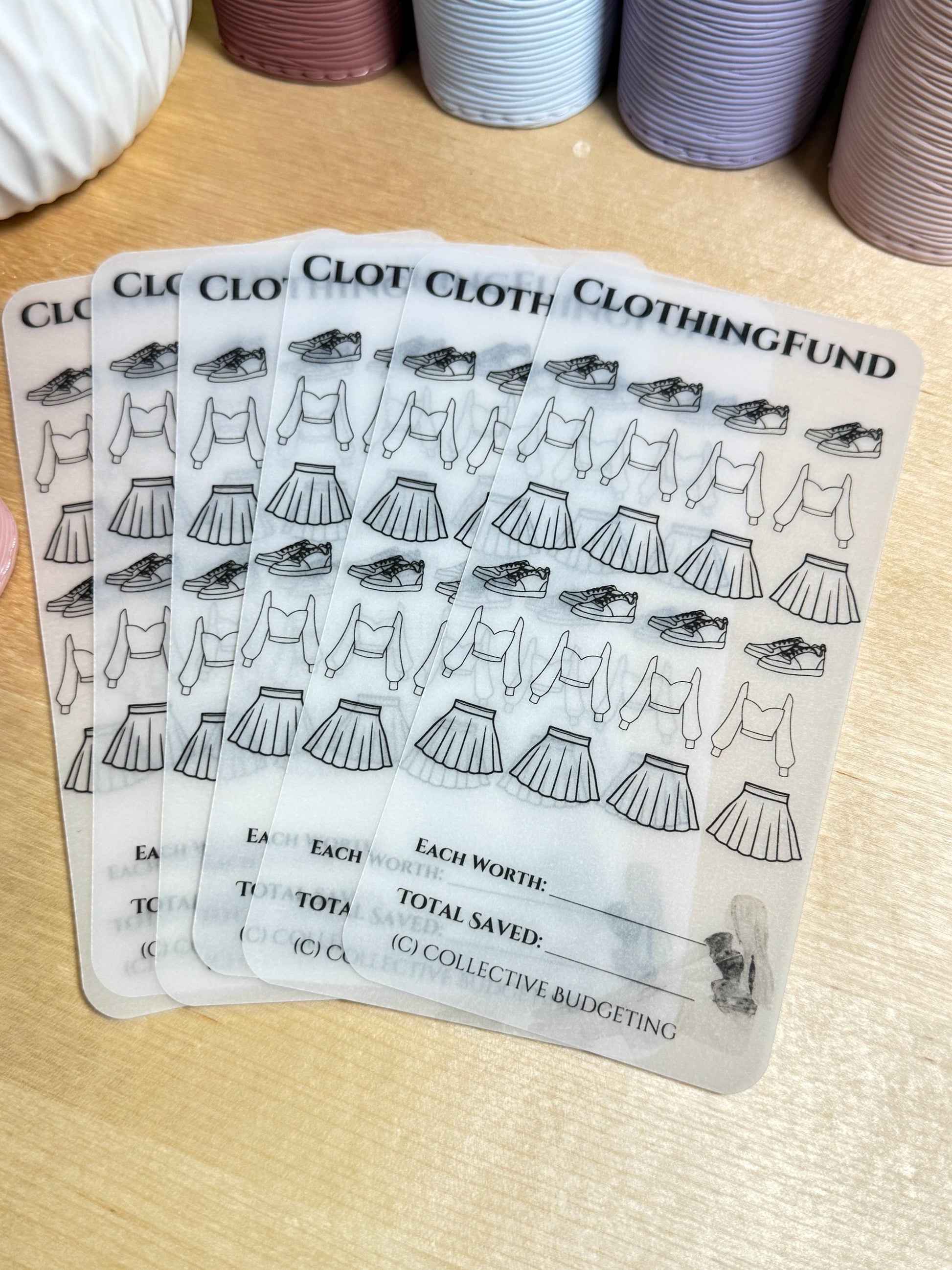 A set of clear plastic sheets with black outlines of clothing items, shoes, and the text 'CLOTHING FUND.' The sheets have sections for writing in the total worth and total saved. The text '(C) COLLECTIVE BUDGETING' is at the bottom of the sheet.