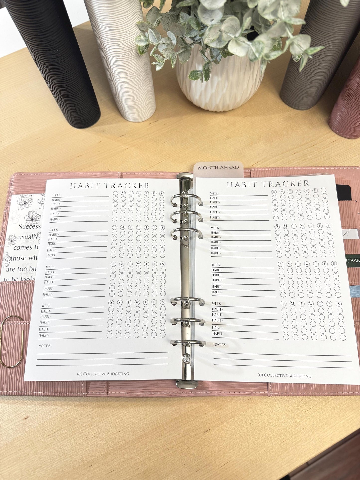 A close-up of a pink planner with two open pages. The pages are habit trackers with a grid of circles for marking habits. The pages are labeled 'Habit Tracker' and 'Month Ahead.' The planner has a silver ring binder and a silver clip.