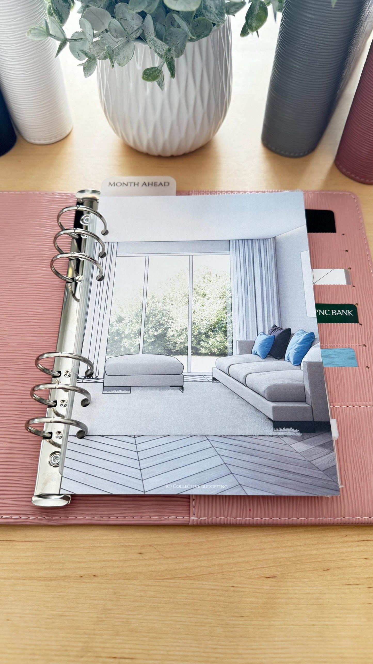 A pink leather bound planner with silver rings. The planner is open to a page with a drawing of a living room with a couch, coffee table, and windows. The page has the text 'MONTH AHEAD' at the top. The planner has a tab that says 'PNC BANK'.