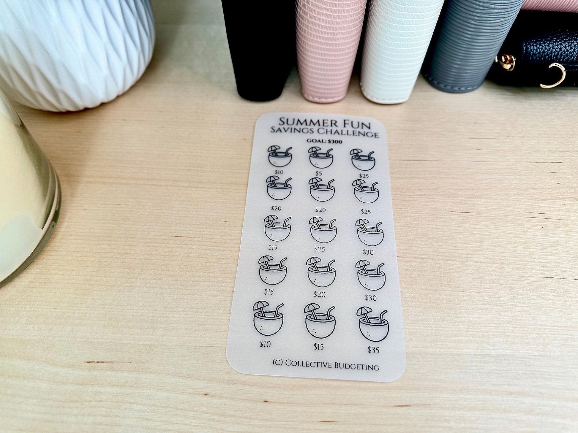 A white sticker with a black outline of a coconut drink with an umbrella and a straw. The sticker is labeled 'Summer Fun Savings Challenge' with a goal of $300. The sticker has 18 spaces to track savings with amounts ranging from $5 to $35.