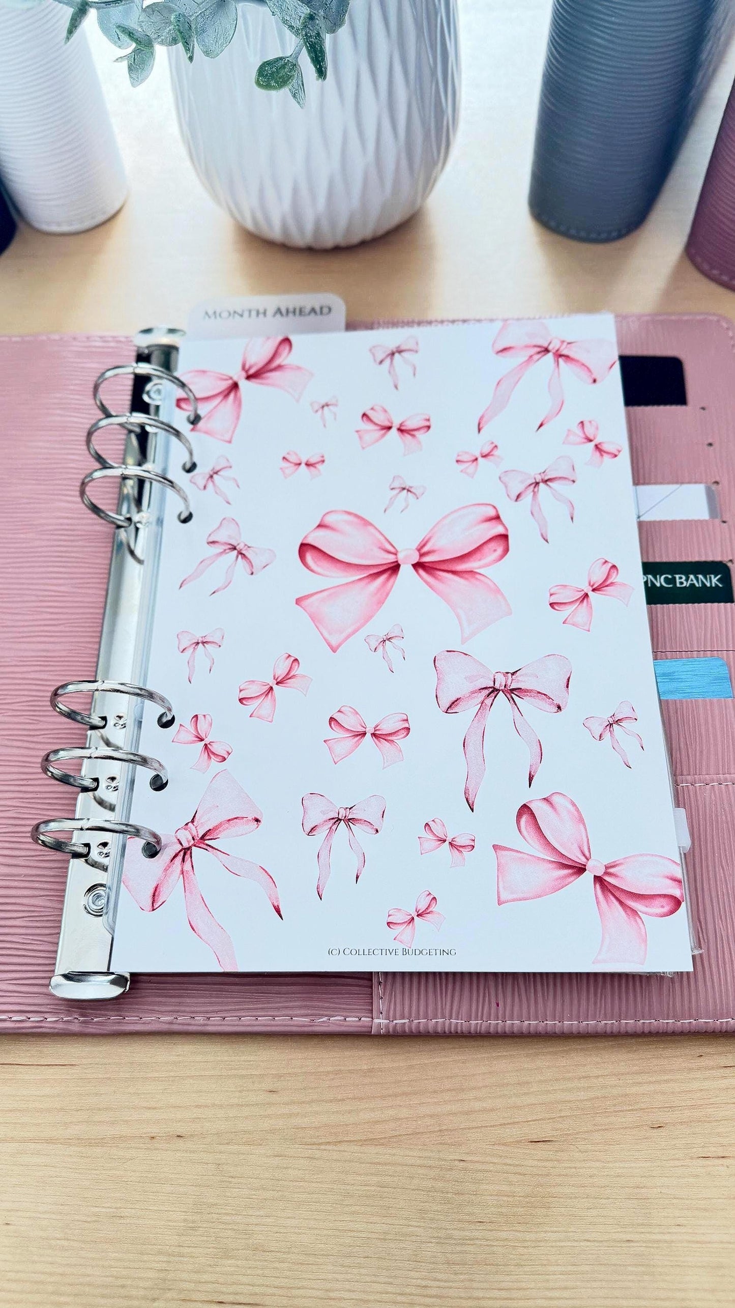 A white planner page with a pink bow pattern. The page has the text 'MONTH AHEAD' and 'IC COLLECTIVE BUDGETING'.